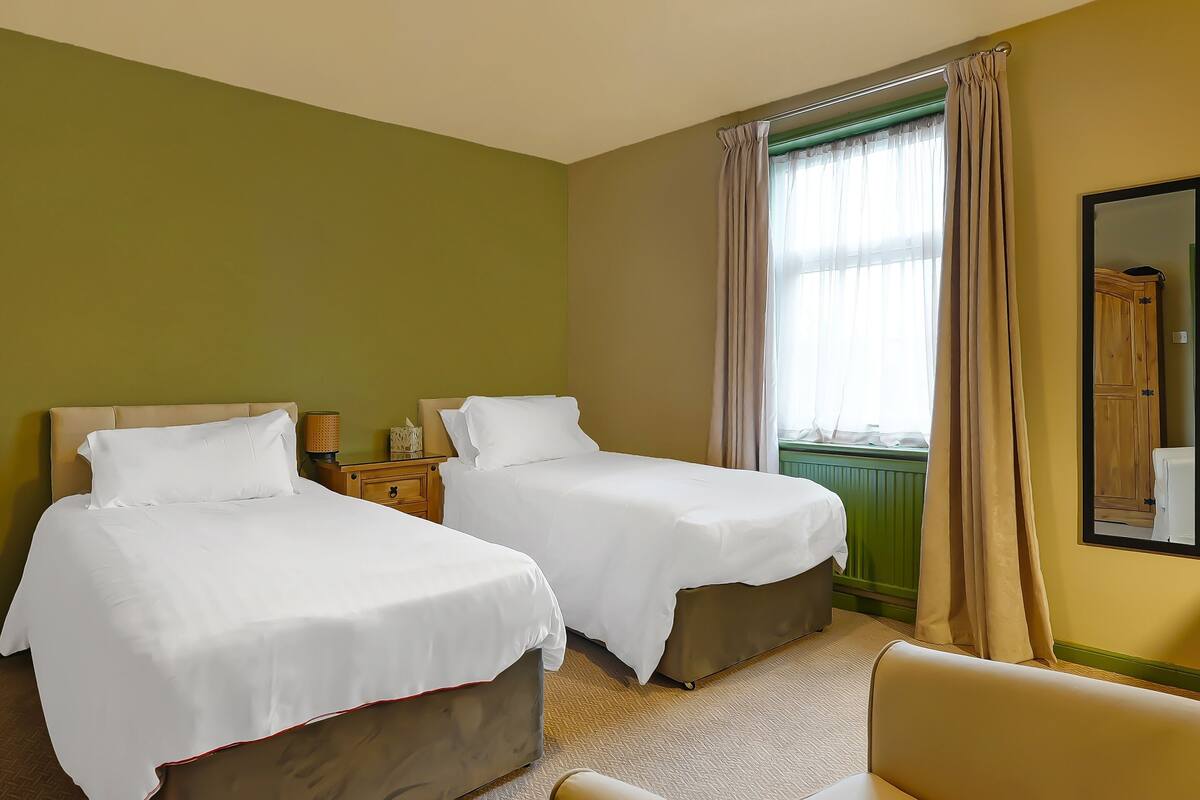 A hotel room with two beds, a chair, a nightstand with a lamp, and a window with curtains.