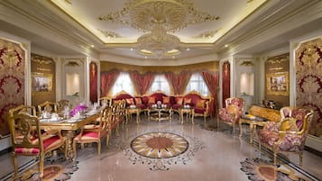 Uma sala de jantar luxuosa com decoração dourada e vermelha elaborada, um grande candelabro e uma mesa redonda central.