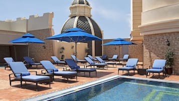 Uma área de piscina com espreguiçadeiras e guarda-sóis, piso revestido e uma estrutura em forma de cúpula ao fundo.