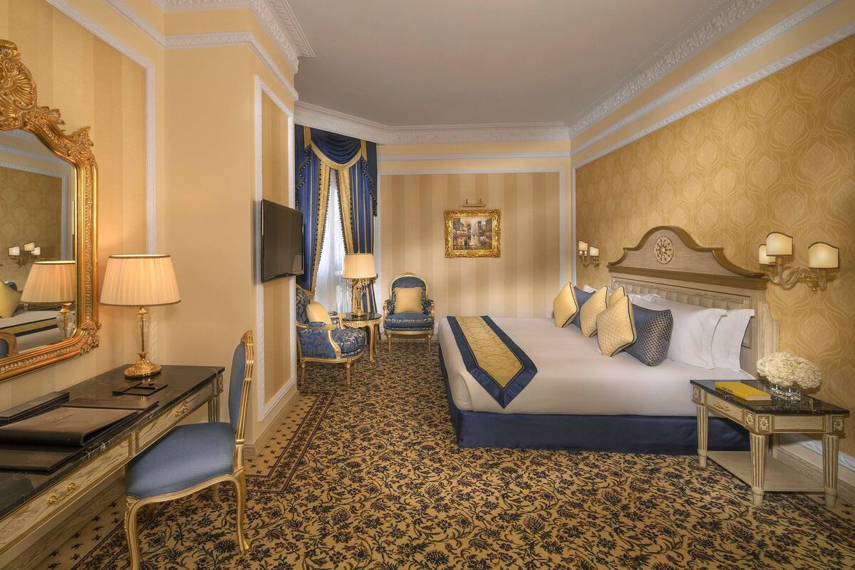 Een luxueuze hotelkamer met een groot bed, een zithoek met een bureau en stoel, een televisie en een grote spiegel.