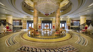 A grand lobby with a large chandelier, ornate columns, and a central seating area with red chairs.