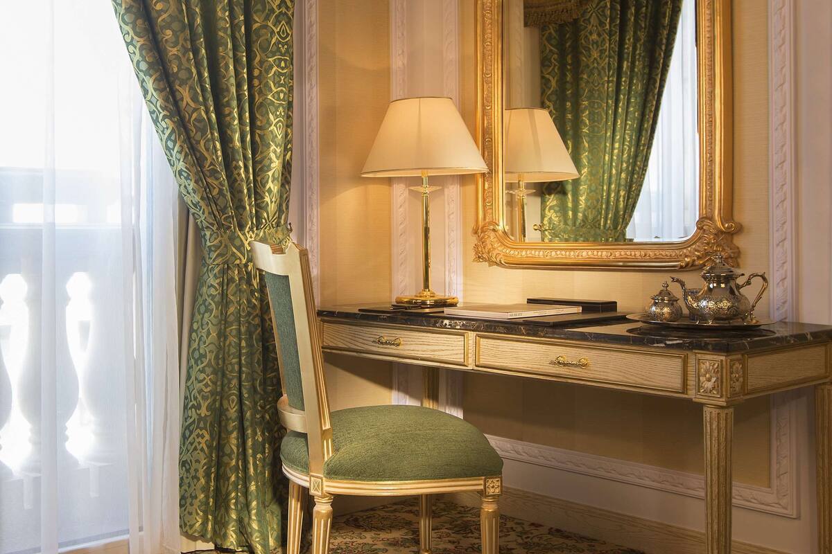 Een kamer met een groen gestreept tapijt, een groene bekleden stoel, een spiegel met gouden lijst en een bureau met een lamp.