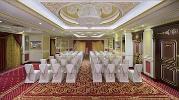 A grand hall with rows of white chairs, a red carpet, and ornate ceiling decorations.