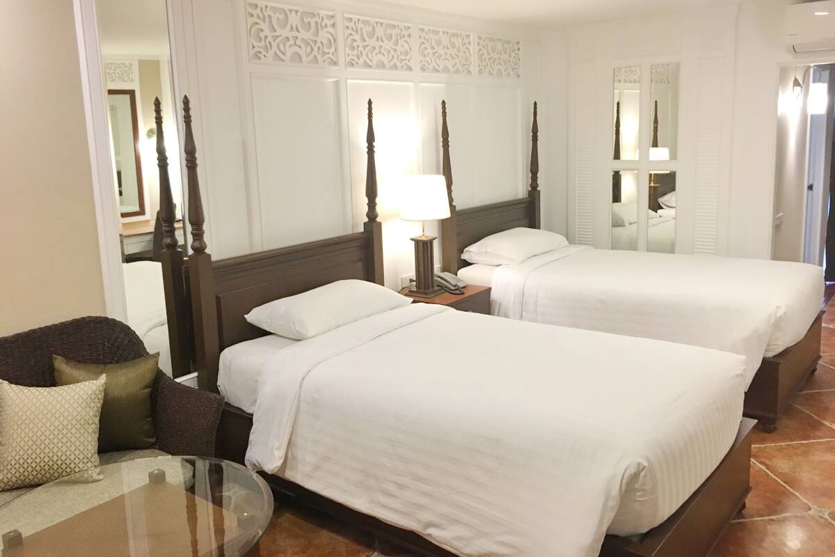 A hotel room with two beds, a nightstand with a lamp, a glass table, and a sofa with cushions.