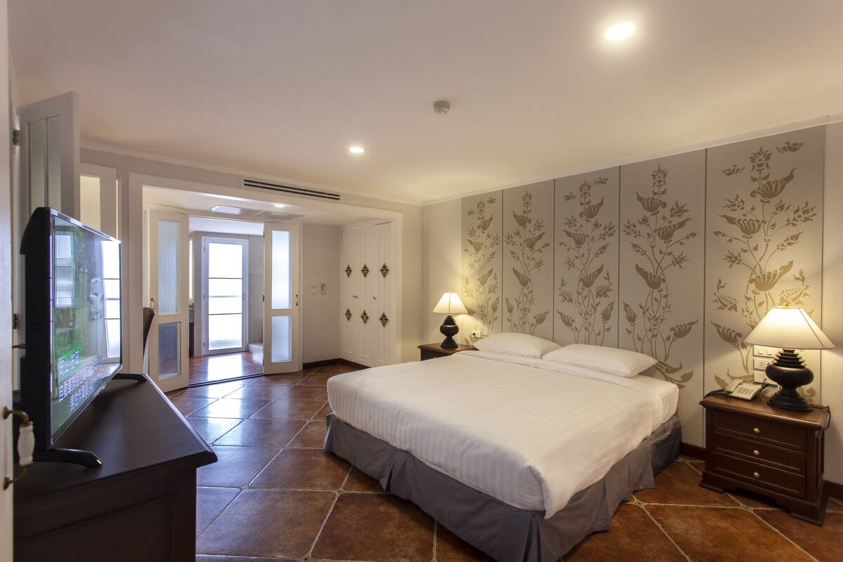 A bedroom with a large bed, a TV on a stand, and a nightstand with a lamp.