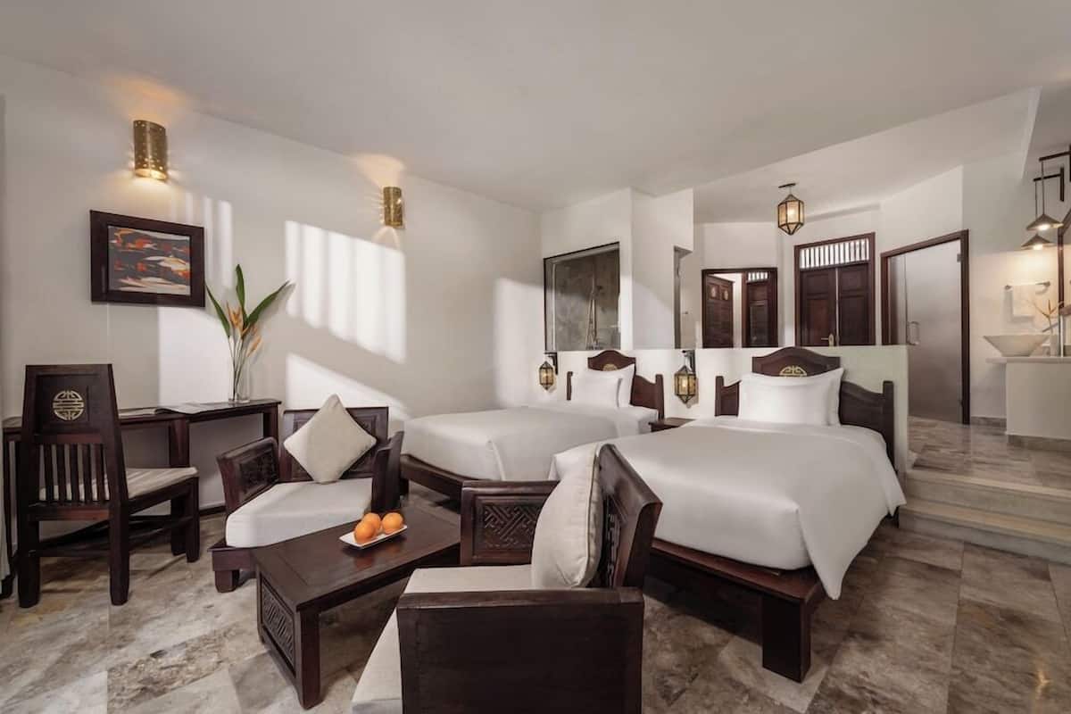 A hotel room with two beds, a desk, a chair, a small table with fruit, and a potted plant.
