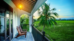 A balcony with wooden chairs, a view of a green field, and a palm tree. - Legacy Hoi An Resort (Da Nang)