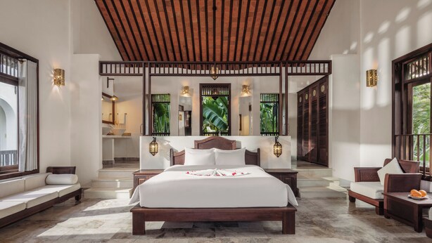 A spacious bedroom with a large bed, a wooden ceiling, and a view of a bathroom and greenery outside. - Legacy Hoi An Resort (Da Nang)