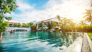 A swimming pool with clear water, surrounded by lush greenery and a building with a red roof. - Legacy Hoi An Resort (Da Nang)