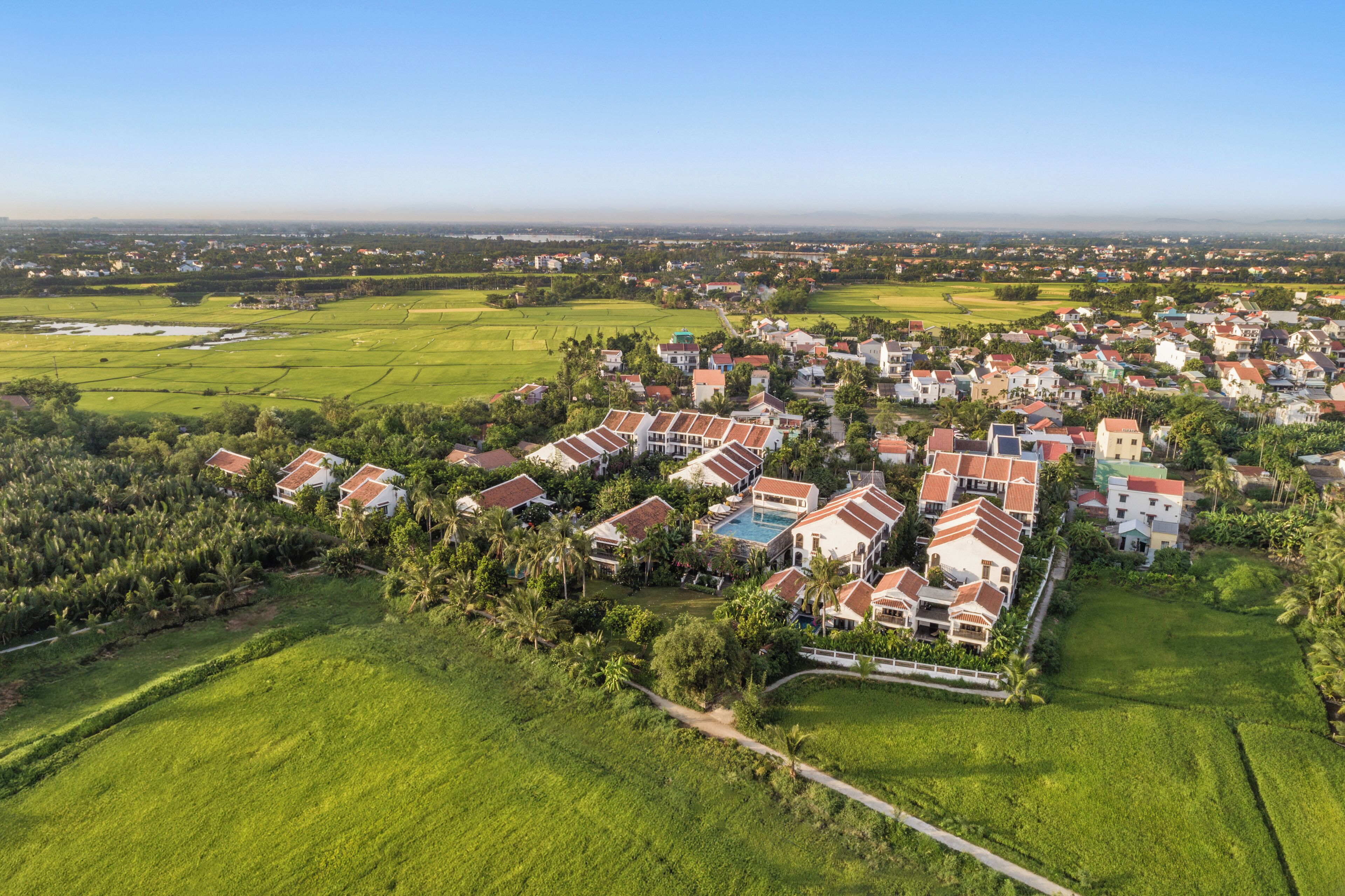 A residential area with houses featuring red roofs and white walls, surrounded by green fields and trees.