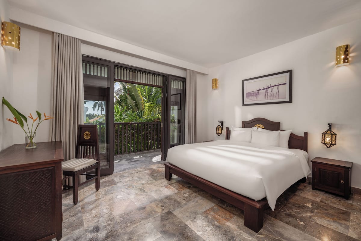 A modern hotel room with a large bed, a desk, and a chair. There is a balcony with a view of greenery.