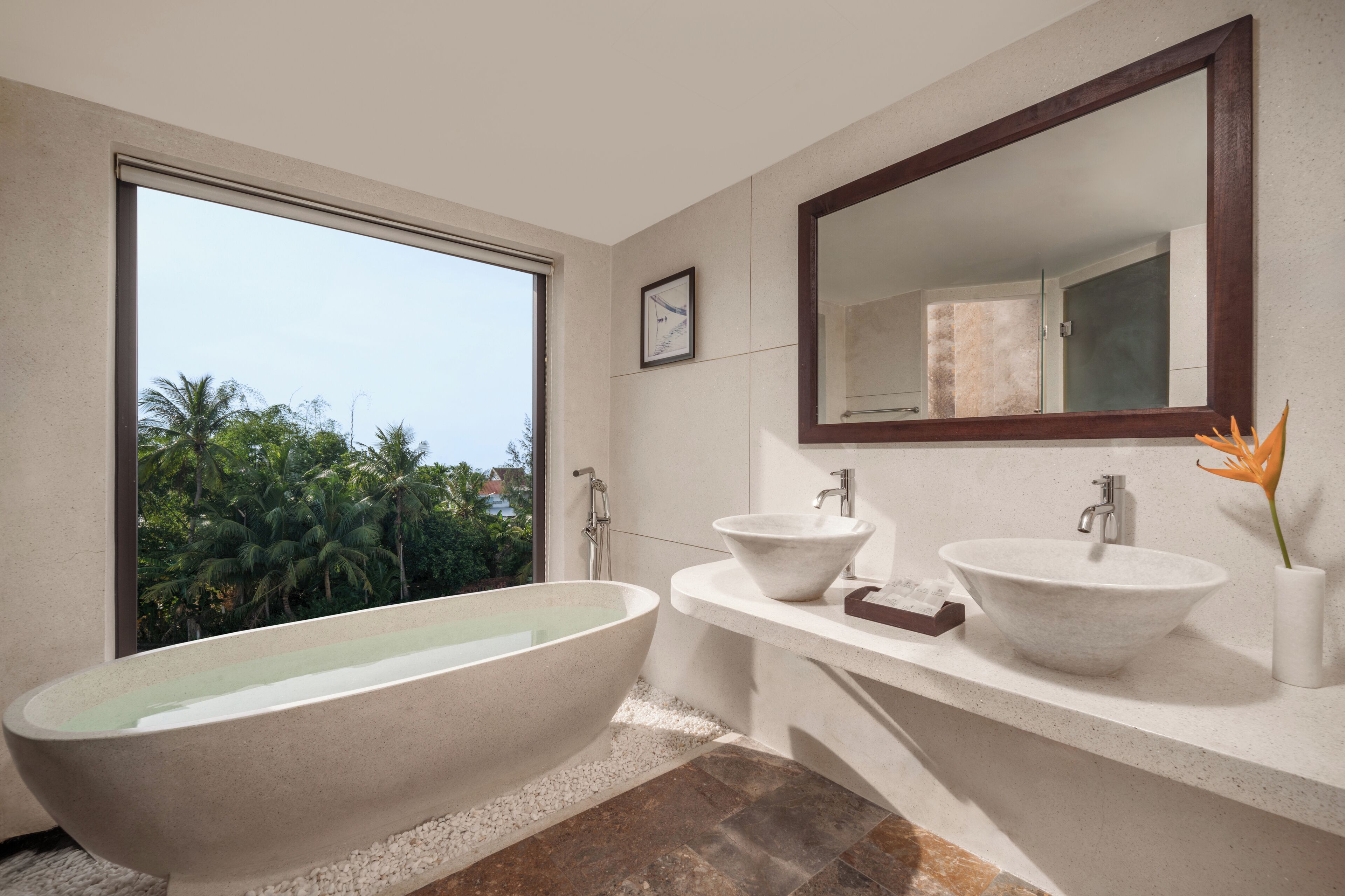 A modern bathroom with a freestanding bathtub, two sinks, a large mirror, and a view of palm trees.