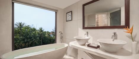 A modern bathroom with a freestanding bathtub, two sinks, a large mirror, and a view of palm trees.