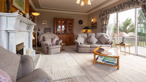A spacious living room with two grey sofas, a white fireplace, and a large window with a view of greenery. - Fern Rock B&B (Milltown)
