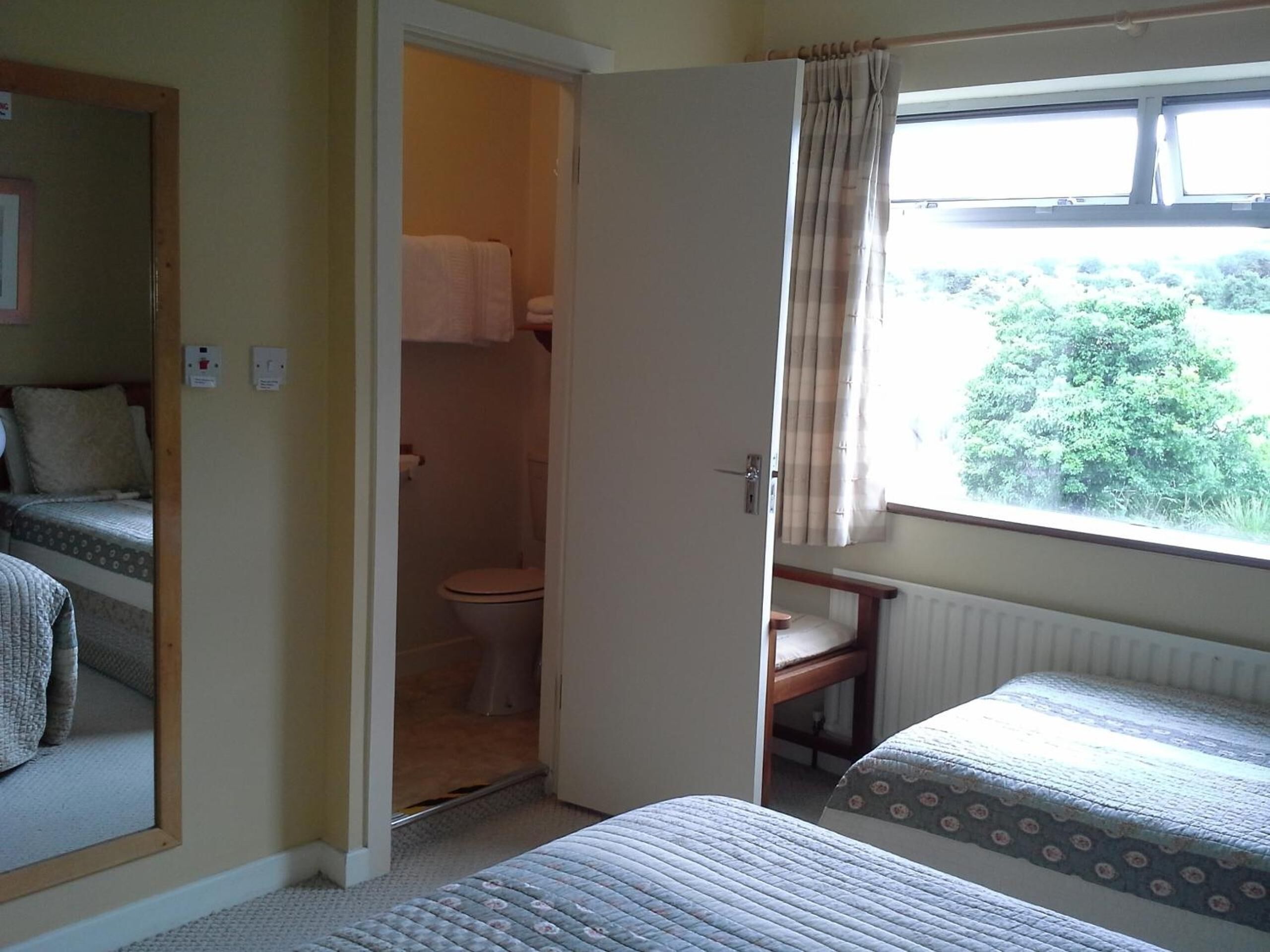 A bedroom with two single beds, a window with curtains, a door leading to a bathroom, and a radiator under the window.