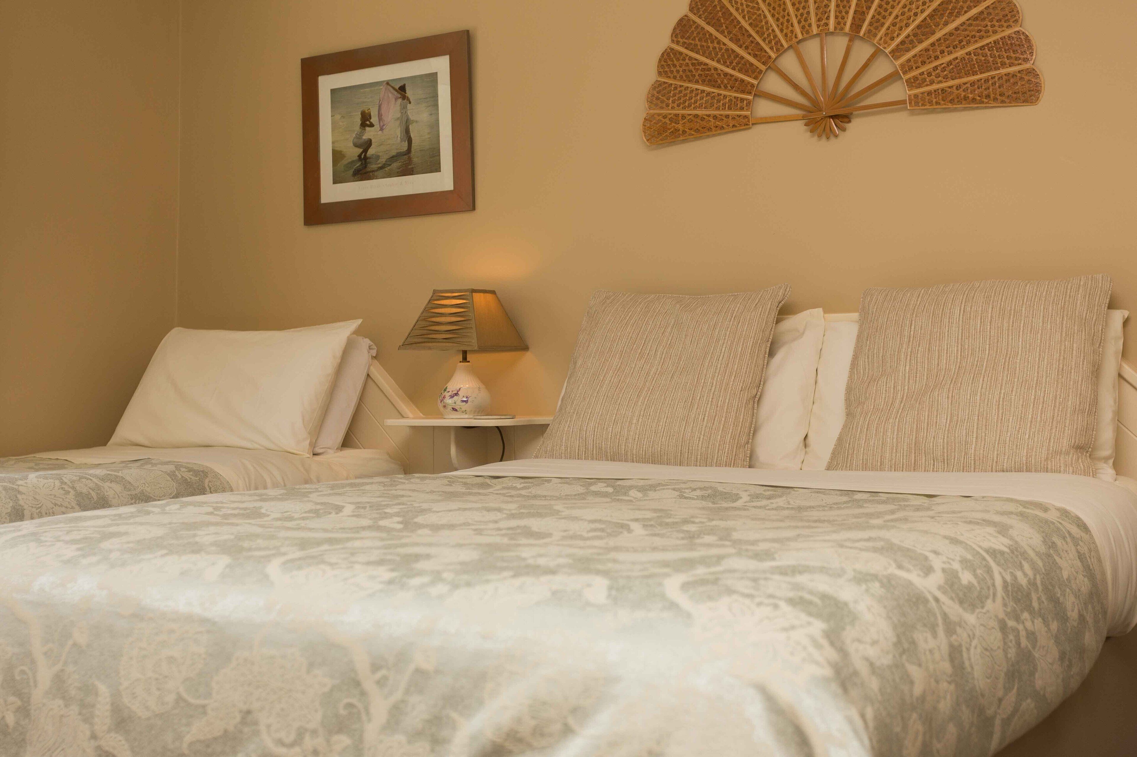 A hotel room with two beds, a bedside table with a lamp, a framed picture on the wall, and a decorative fan.
