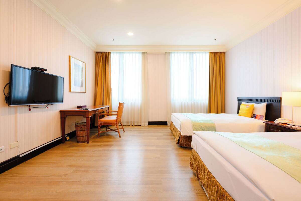 Ein Hotelzimmer mit zwei Betten, einem Schreibtisch mit Stuhl, einem Fernseher und einem Fenster mit Vorhängen.