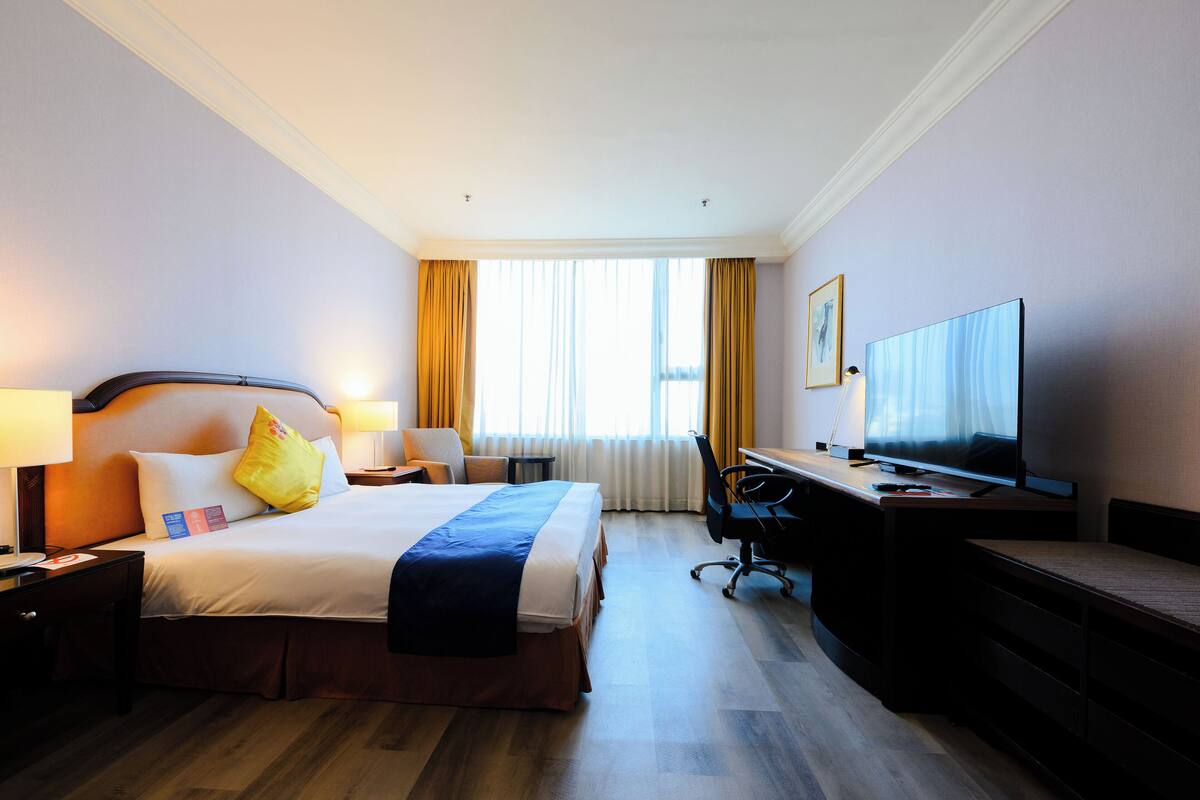 Ein Hotelzimmer mit einem großen Bett, einem Schreibtisch mit Computer, einem Fernseher und einem Fenster mit Vorhängen.