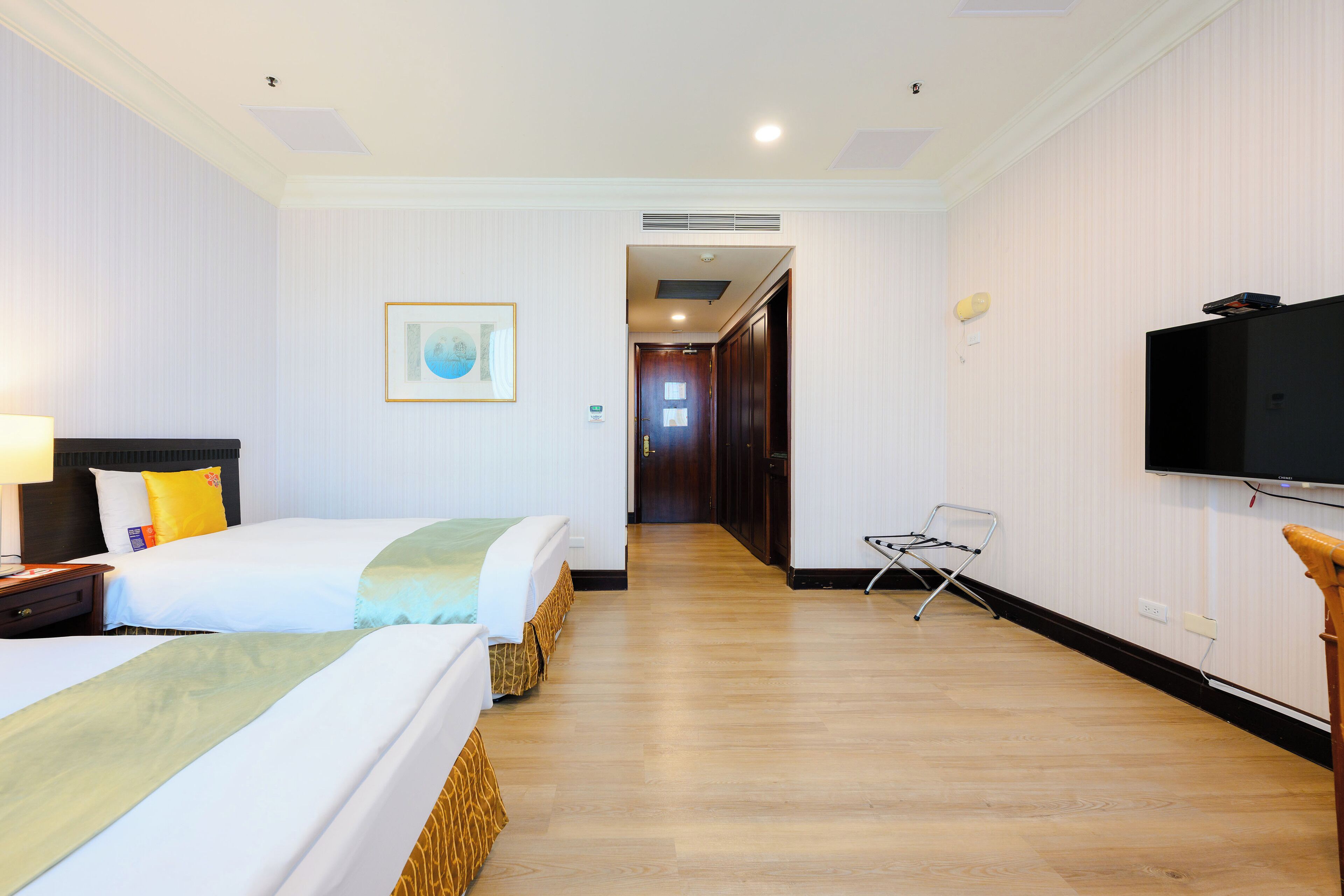 A hotel room with two beds, a television, a chair, and a hallway leading to another room.