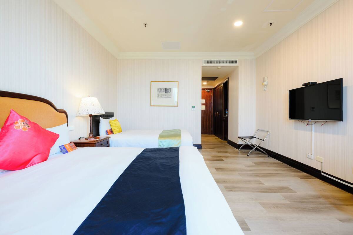 Ein Hotelzimmer mit zwei Betten, einem Fernseher, einer Lampe und einem gerahmten Bild an der Wand.