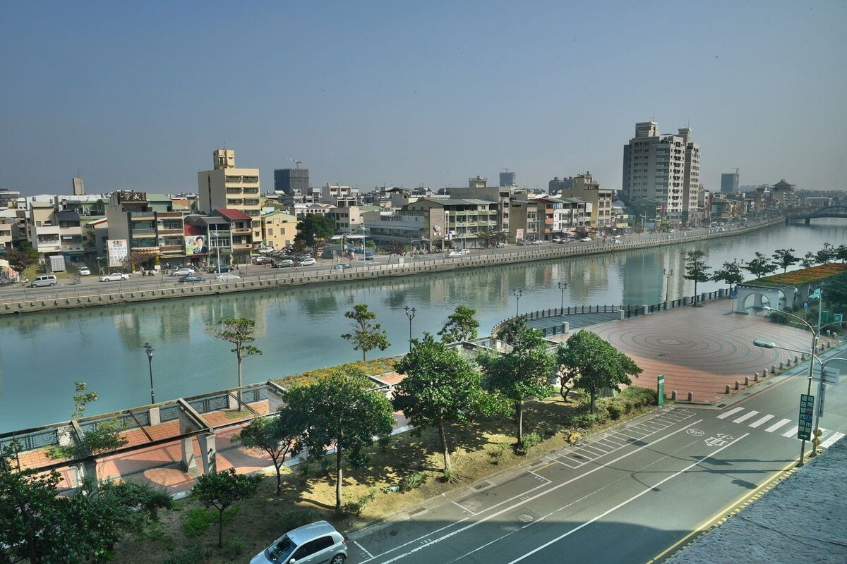 A riverside cityscape with buildings, a pedestrian walkway, and a parked car.