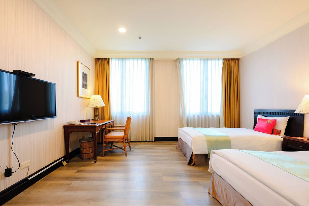 Ein Hotelzimmer mit Flachbildfernseher, Schreibtisch mit Stuhl, zwei Betten mit Kissen und einem Fenster mit Vorhängen.