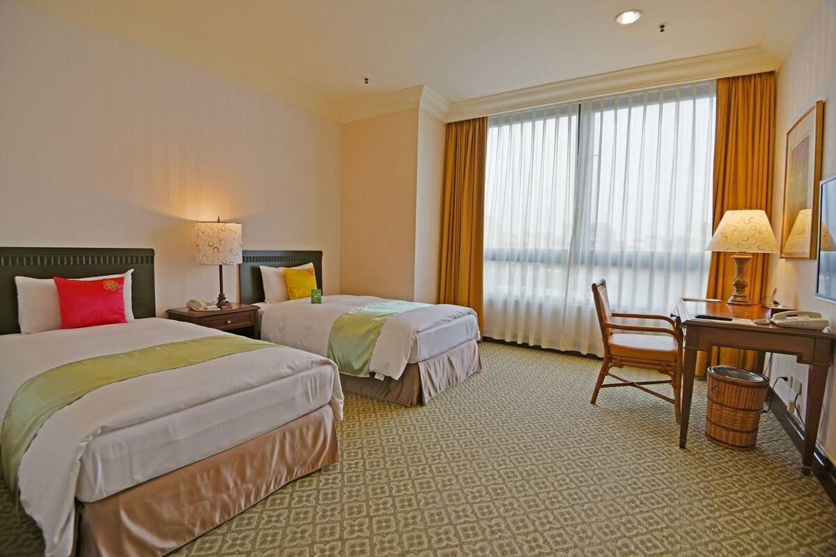 Ein Hotelzimmer mit zwei Betten, einem Schreibtisch, einem Stuhl, einem Mülleimer und einem großen Fenster mit Vorhängen.