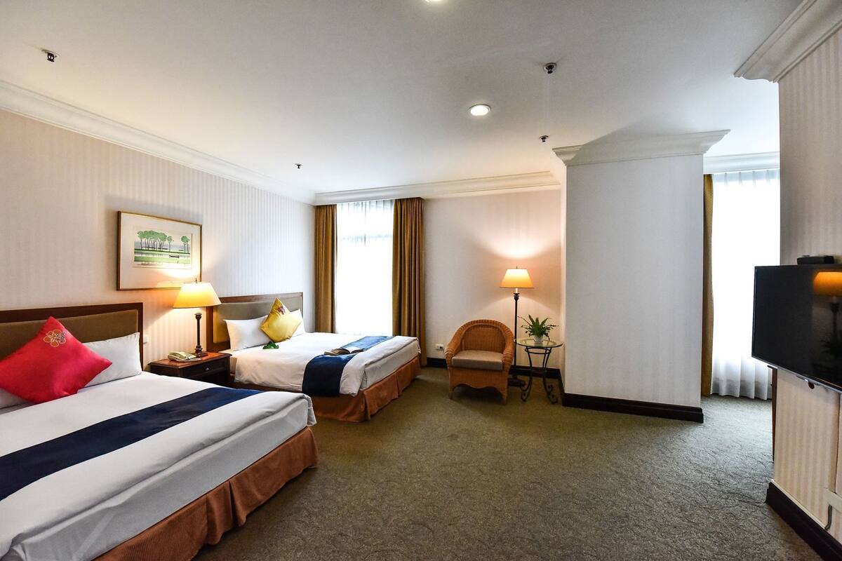 Ein Hotelzimmer mit zwei Betten, einem Sessel, einer Lampe, einem Fernseher und einem gerahmten Bild an der Wand.