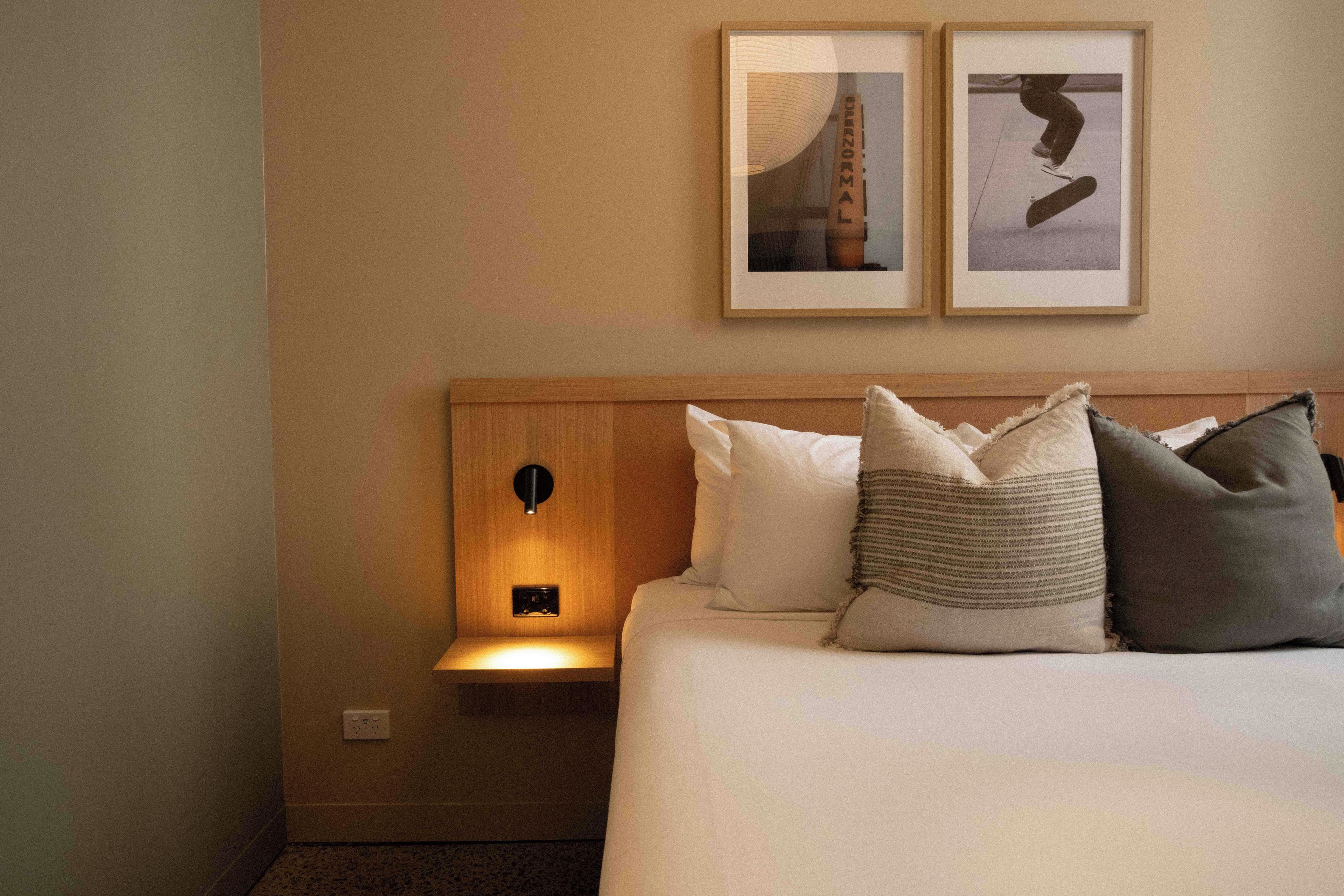A hotel room with a bed, pillows, a nightstand, a wall-mounted lamp, and two framed pictures.