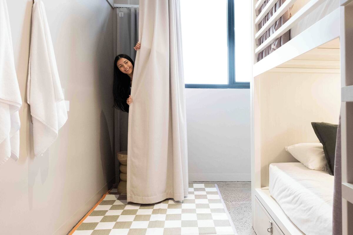 A narrow hallway with a checkered floor, a bed with a white mattress and pillows, and a window with curtains.