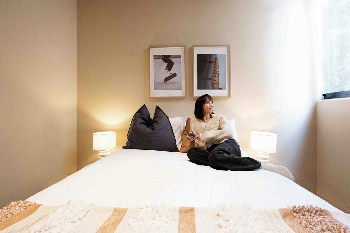 A person sitting on a bed in a hotel room with two framed pictures on the wall, two bedside lamps, and a window with curtains.