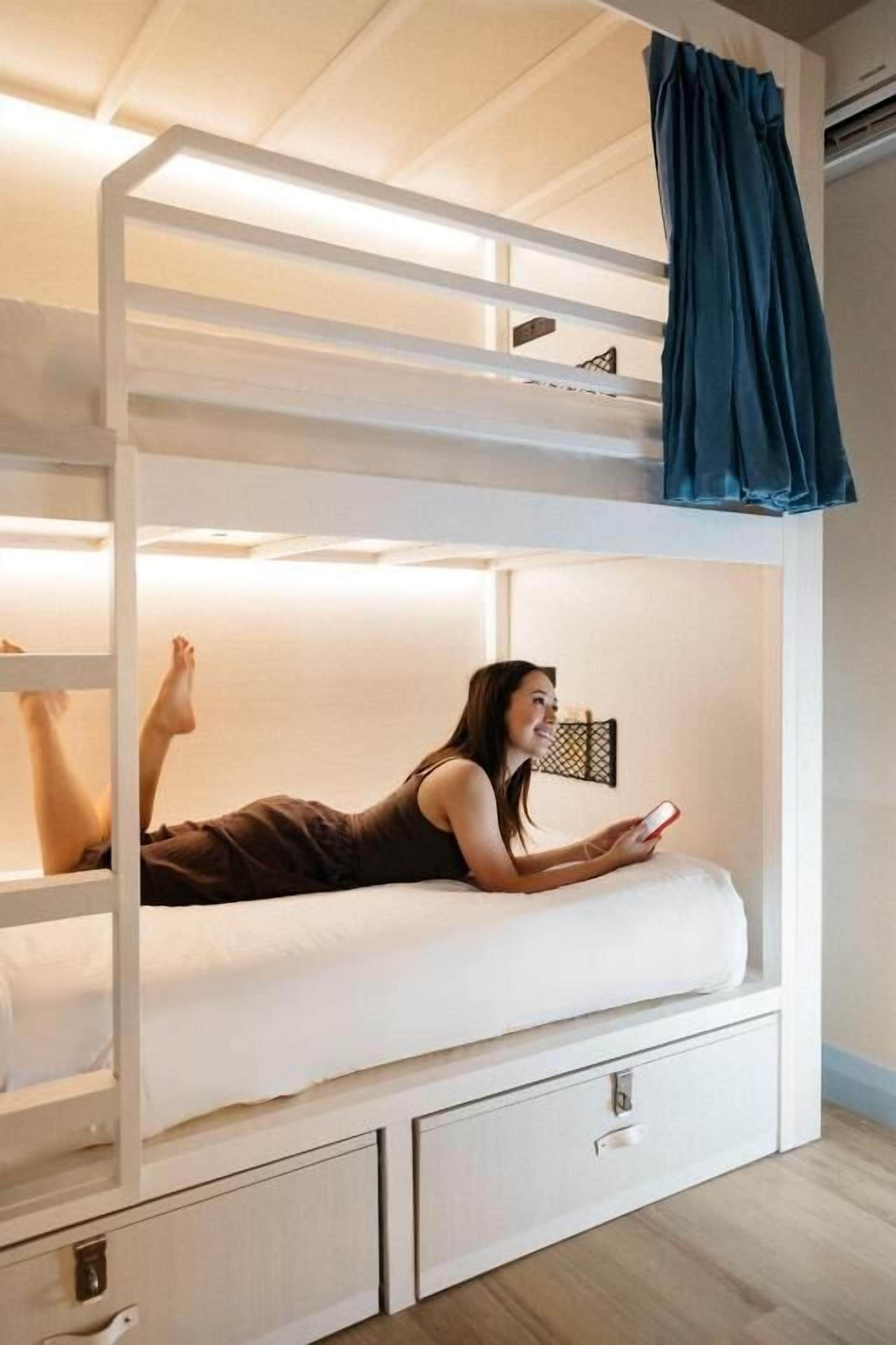A bunk bed with a person lying on the lower bunk, a storage drawer below, and a curtain on the upper bunk.