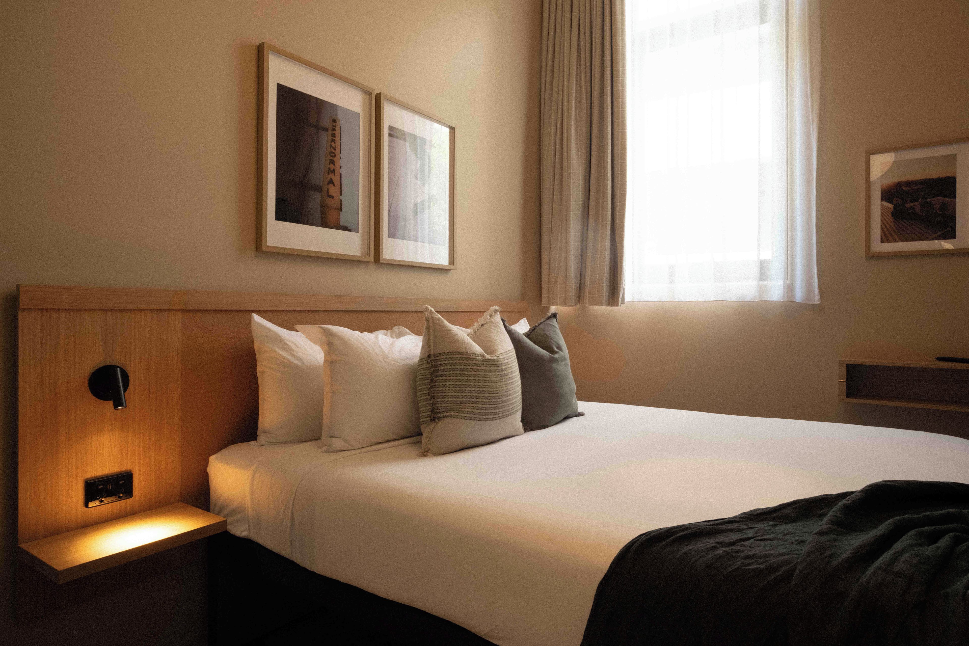 Ein Hotelzimmer mit einem Bett, Kissen, einem Nachttisch, einer Wandleuchte und gerahmten Bildern.