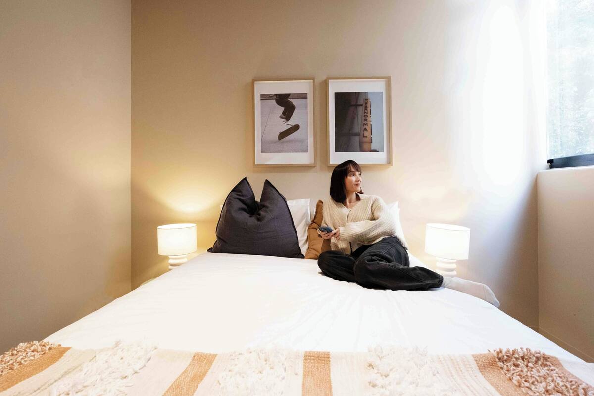 A person sitting on a bed in a hotel room with two framed pictures on the wall, two bedside lamps, and a window with curtains.