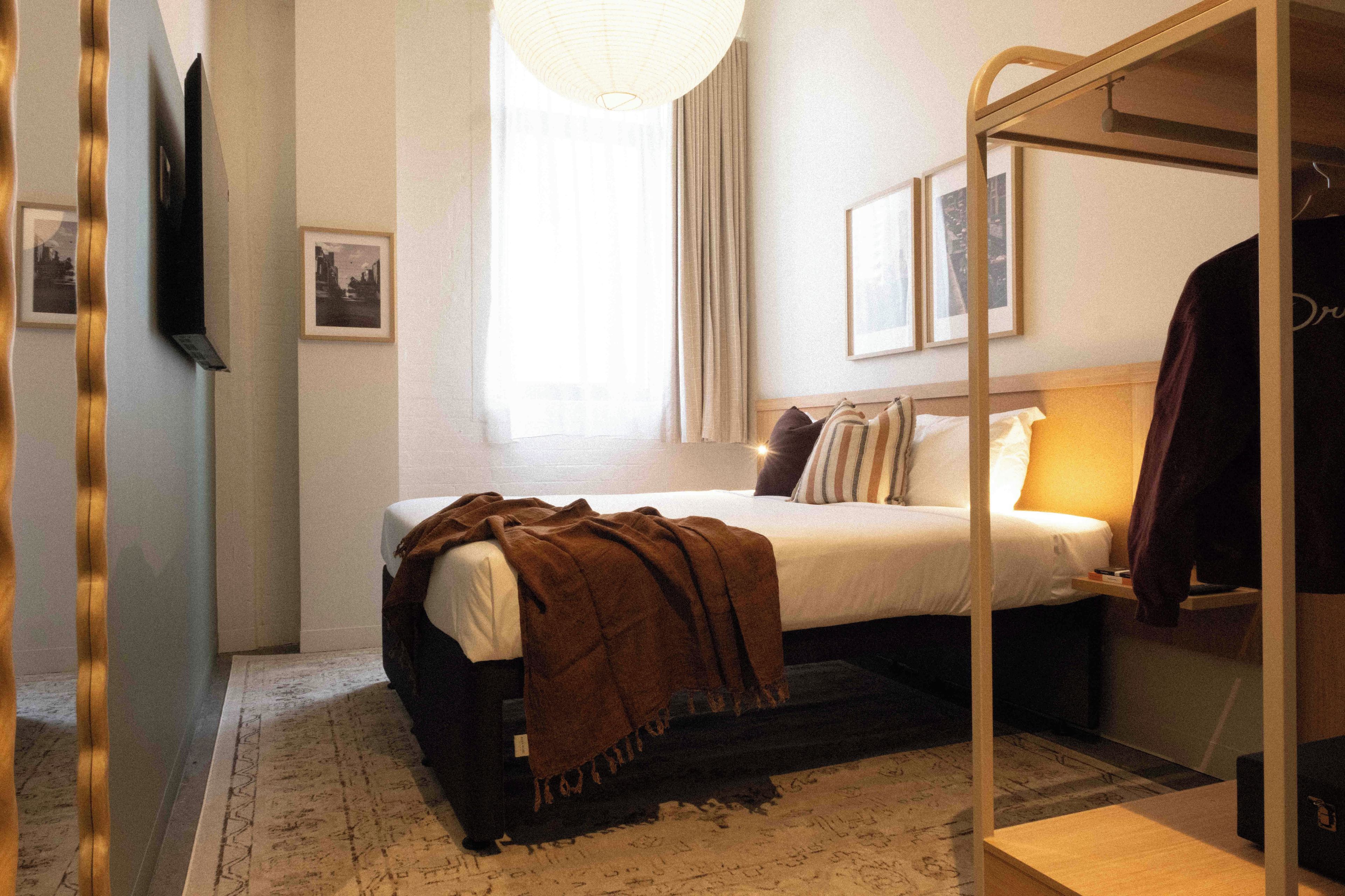 A bedroom with a bunk bed, a bedside table, a lamp, a hanging light fixture, a window with curtains, and a wall-mounted TV.
