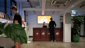 Una zona de recepción con un mostrador de madera, una pantalla grande mostrando información sobre eventos y una planta en maceta.