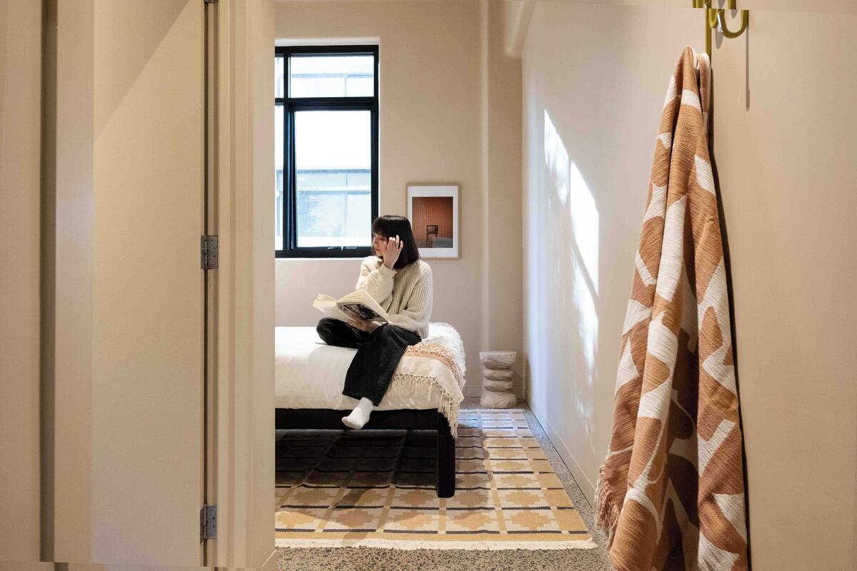 A person sitting on a bed in a hallway, reading a book. There is a window with a view of the outside, a framed picture on the wall, and a patterned rug on the floor.