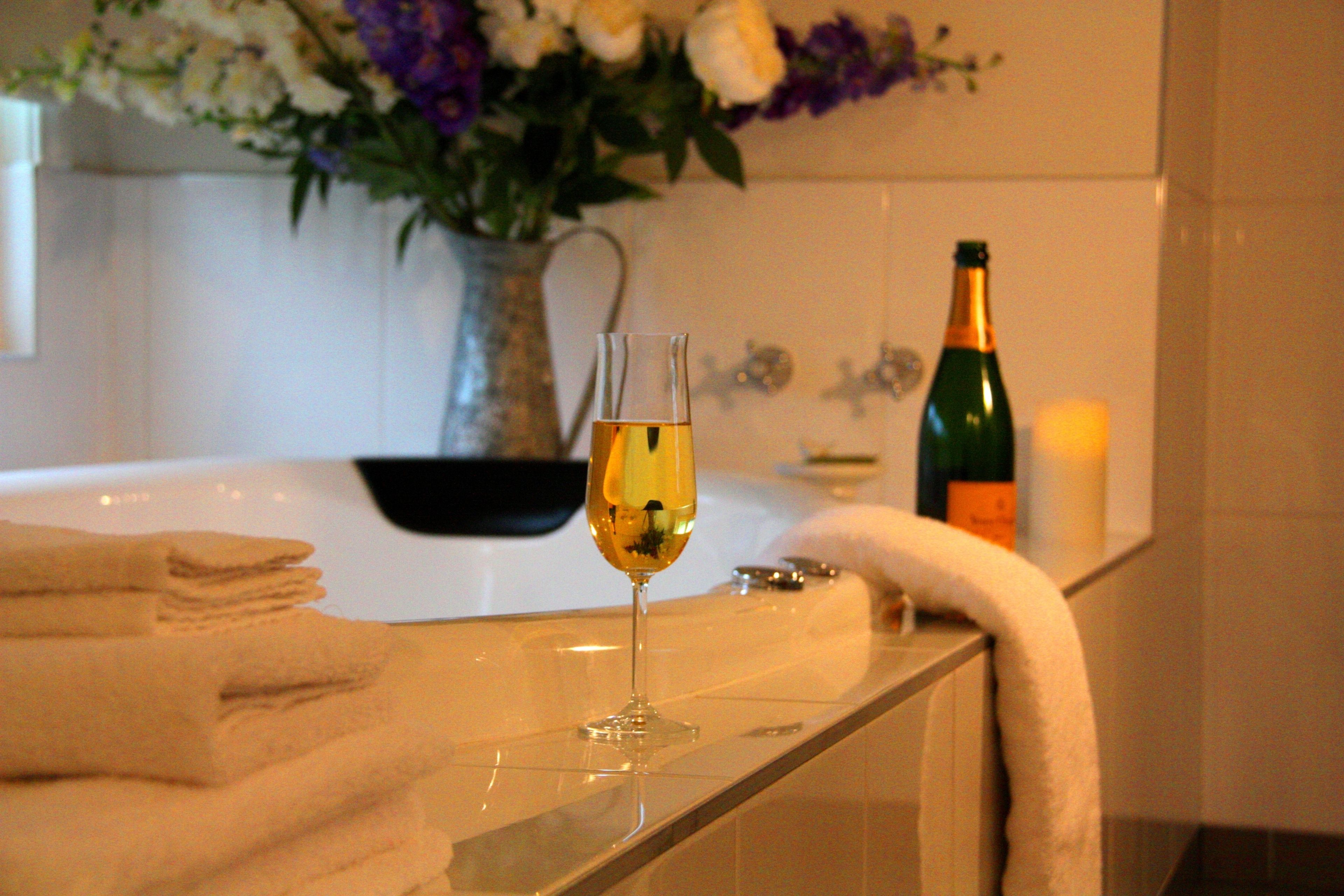 Een badkamer met een glas witte wijn, een kaars en een bloemenvaas op het aanrecht.