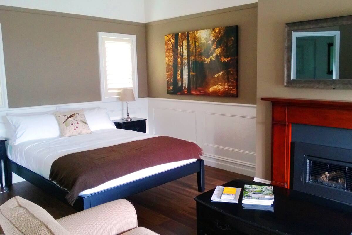 A bedroom with a bed, bedside table, chair, fireplace, and a painting on the wall.