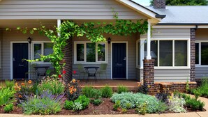 A house with a covered porch, a brick chimney, and a garden with various plants and flowers. - Marysville Garden Cottages (Marysville)