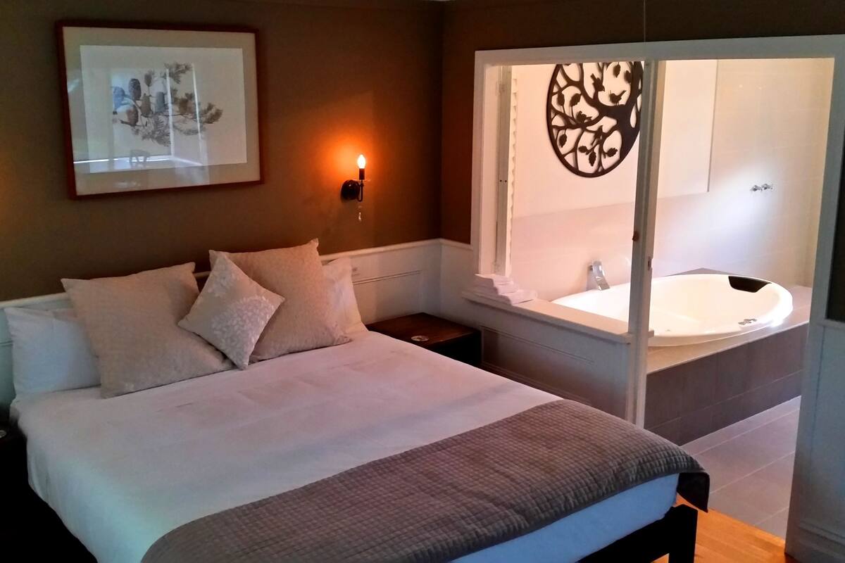 A bedroom with a bed, bedside lamps, a framed picture, and a window with a view of a bathroom.