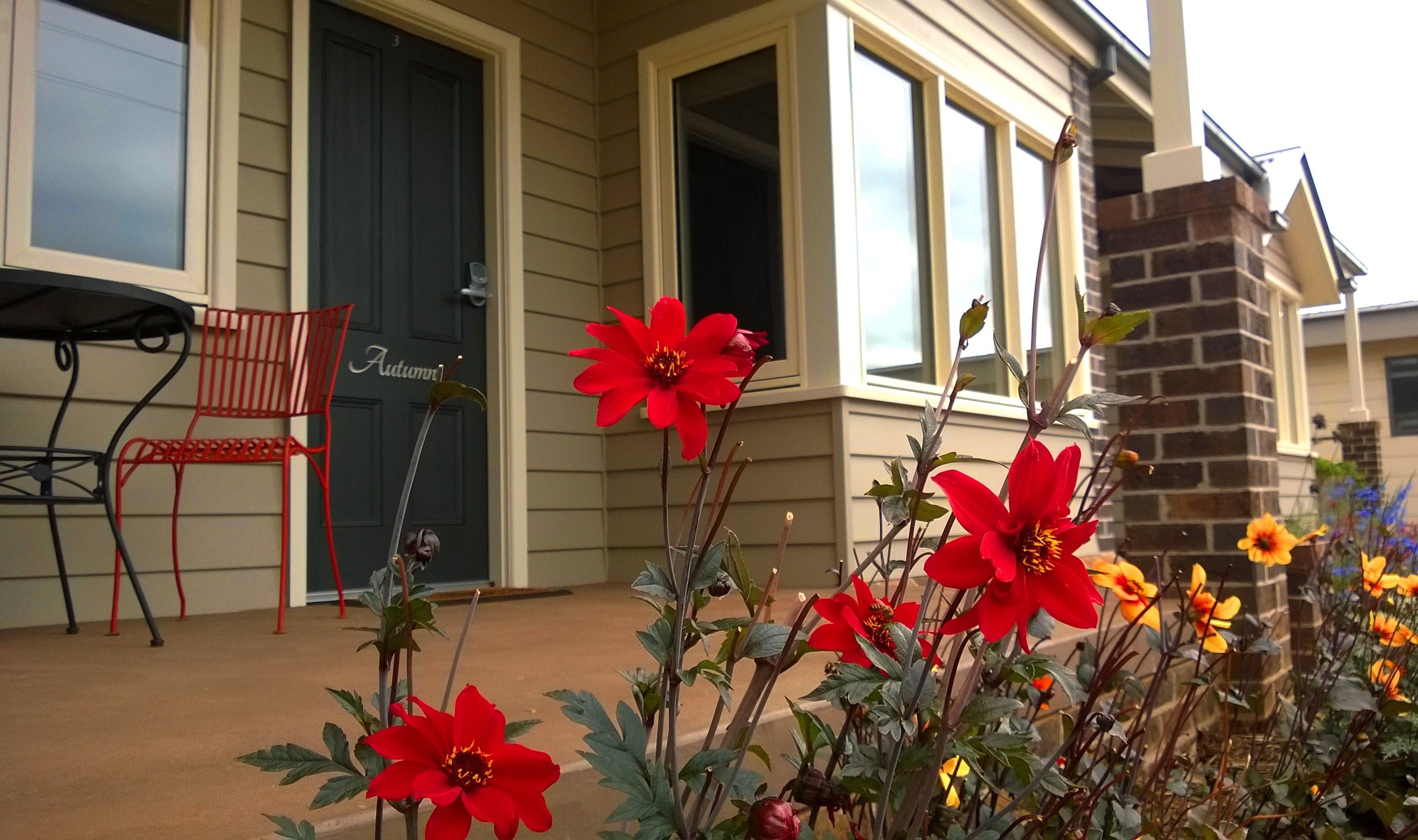 Una casa con puerta principal oscura, una silla roja en el exterior y diversas flores en el jardín delantero.