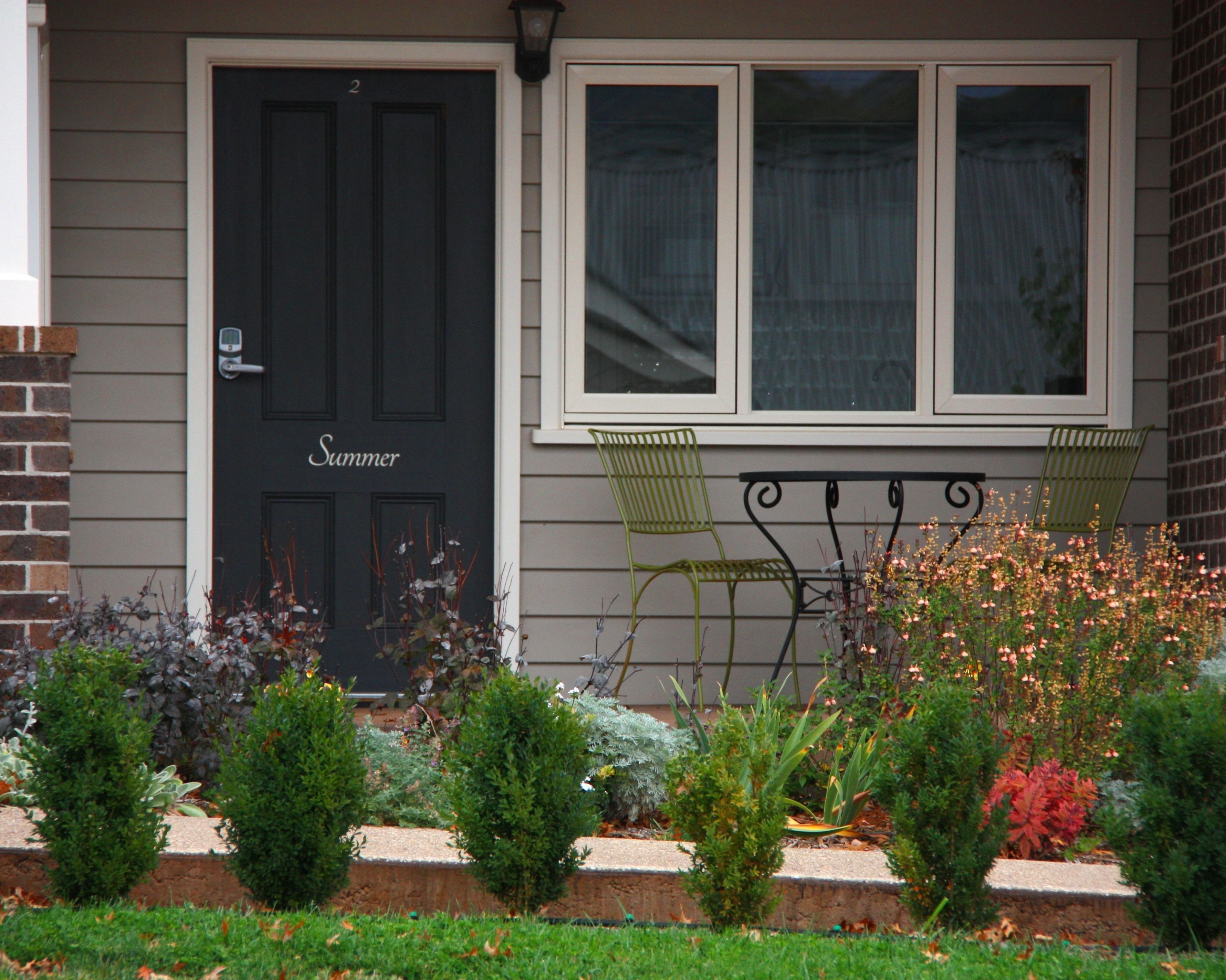 A house with a black door labeled "Summer," a window with white curtains, and a small garden with various plants and a bench.