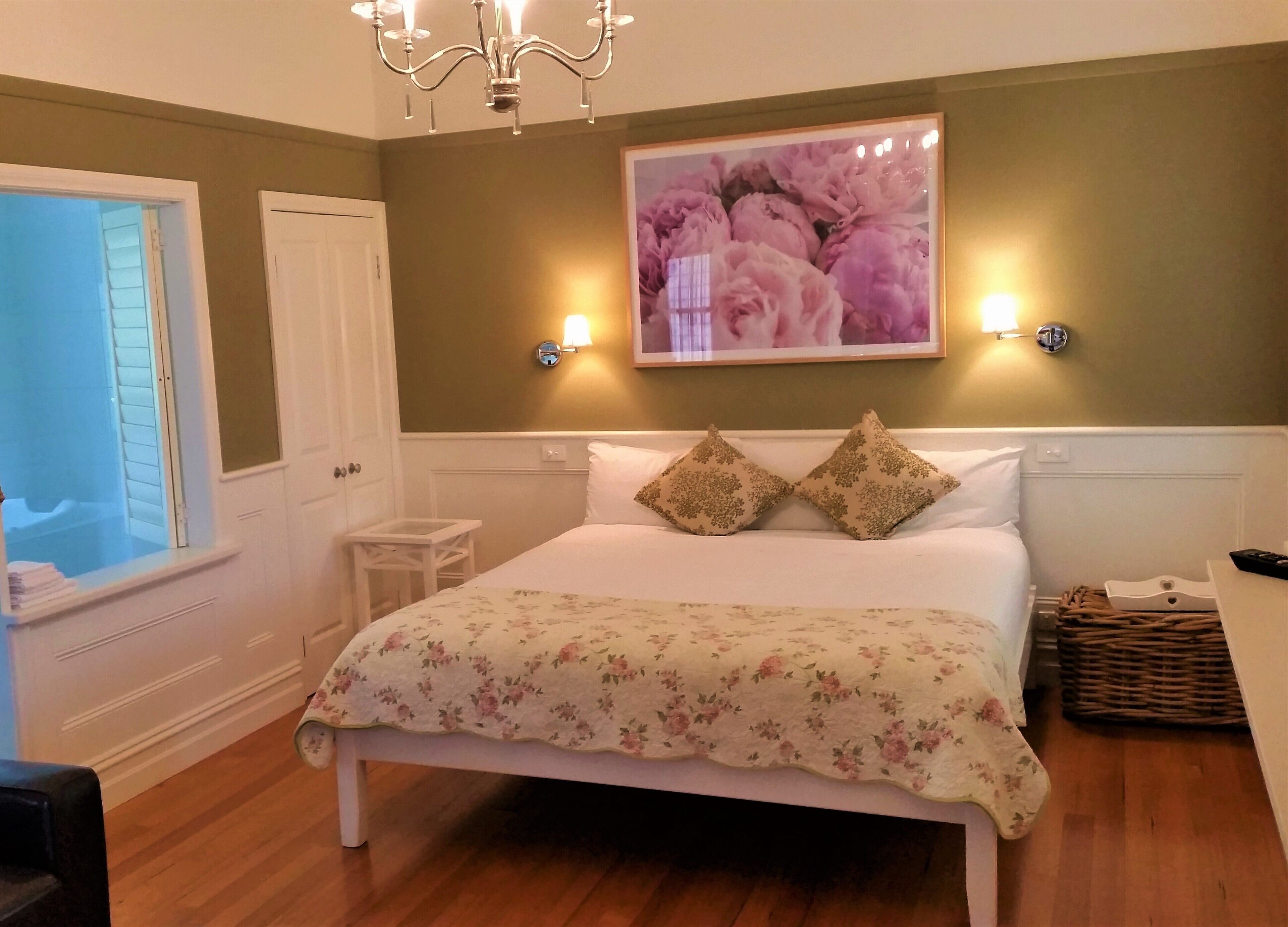A bedroom with a bed, a chandelier, a framed floral picture, and a window.