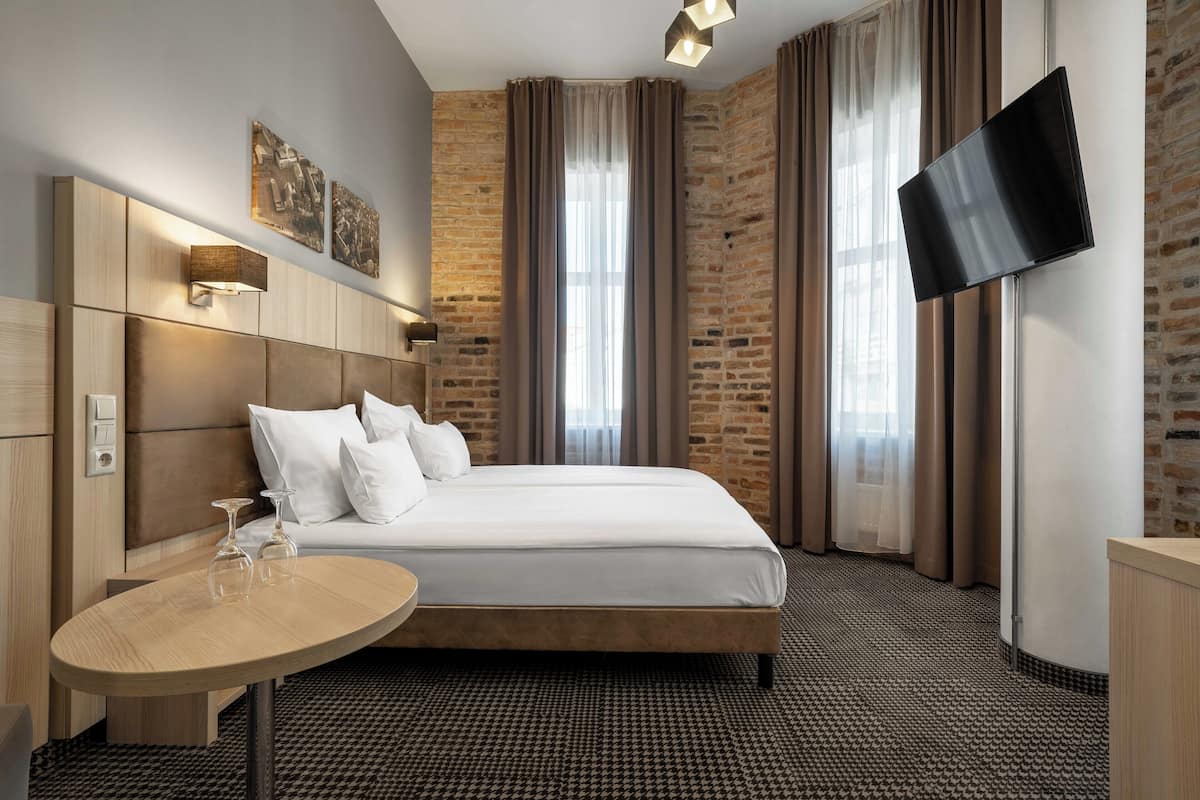 Hotellihuone, jossa on suuri sänky, yöpöytä, jossa on kaksi lasia, seinälle asennettu taulutelevisio ja tiilikuviolla koristeltu seinä.