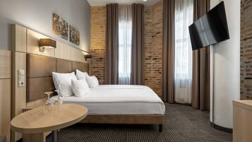 A hotel room with a large bed, a bedside table with two glasses, a flat-screen TV mounted on the wall, and a brick-patterned wall.