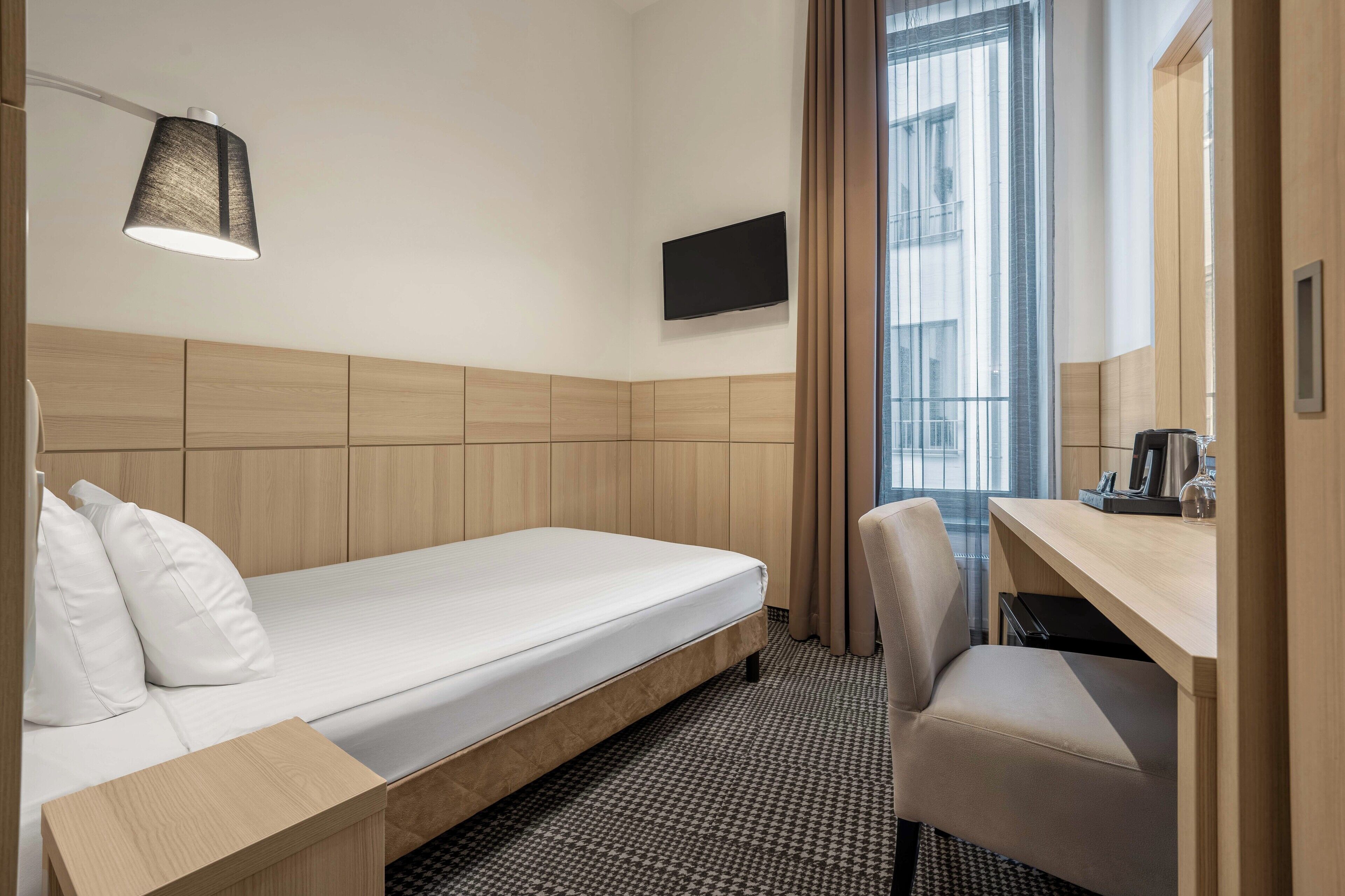 Habitación de hotel con una cama individual, un escritorio con silla, un televisor de pantalla plana en la pared y una ventana con cortinas.