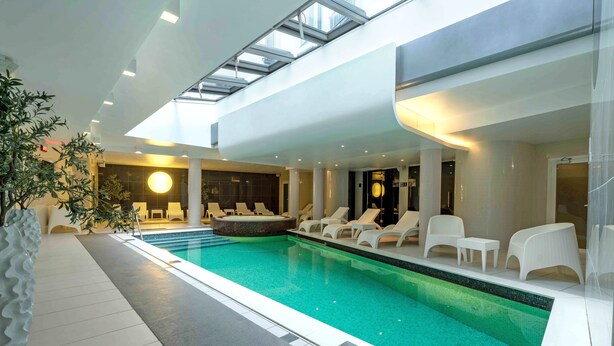 Una piscina interior con sillas de descanso y zona de hidromasaje, rodeada de arquitectura moderna y luz natural.
