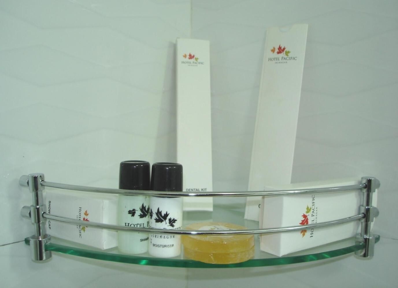 A glass shower tray with a metal frame, containing a yellow soap bar, two bottles of Hotel Pacific shampoo, and a dental kit.