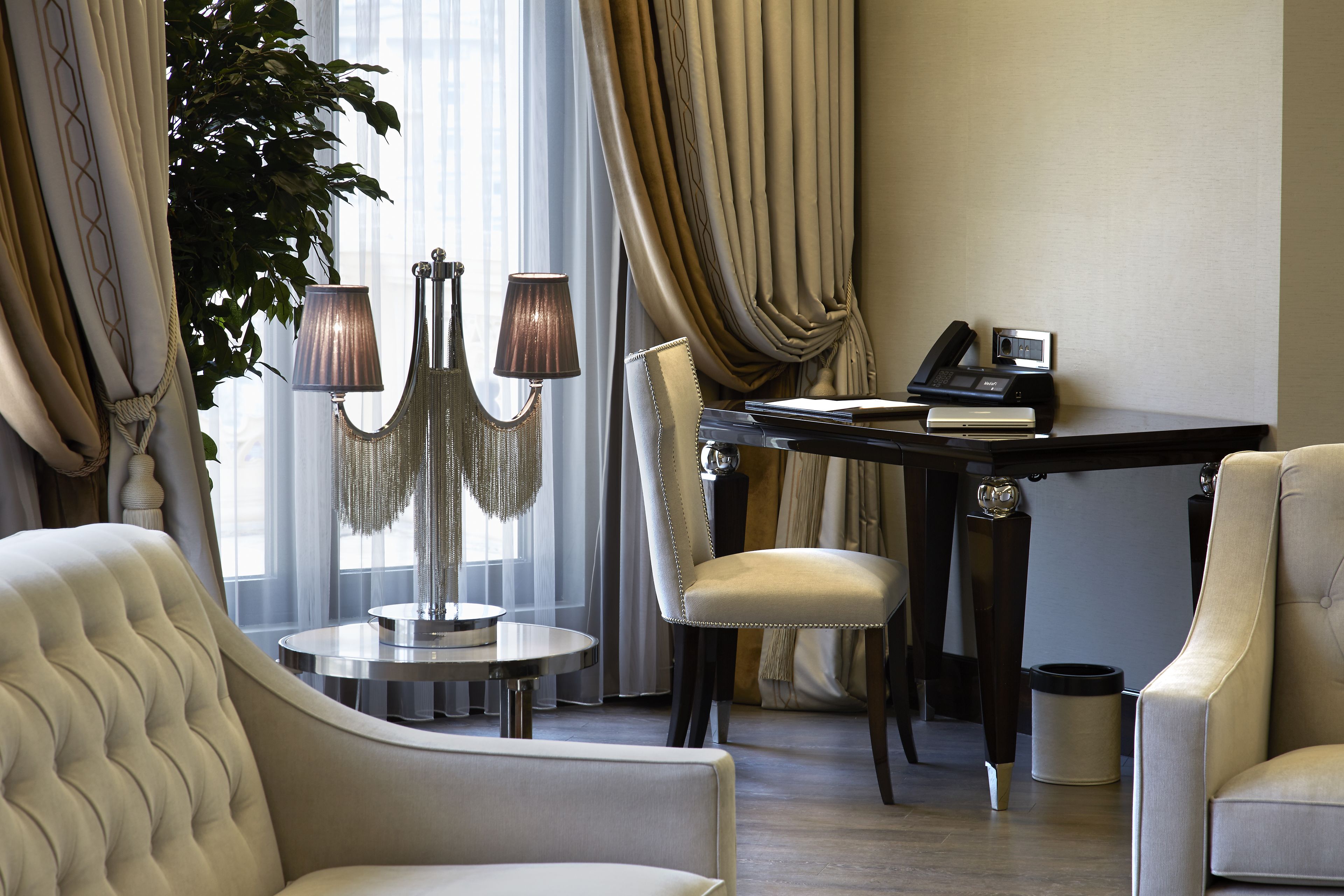 A hotel room with a tufted armchair, a round table with a lamp, a desk with a telephone, and a large plant.